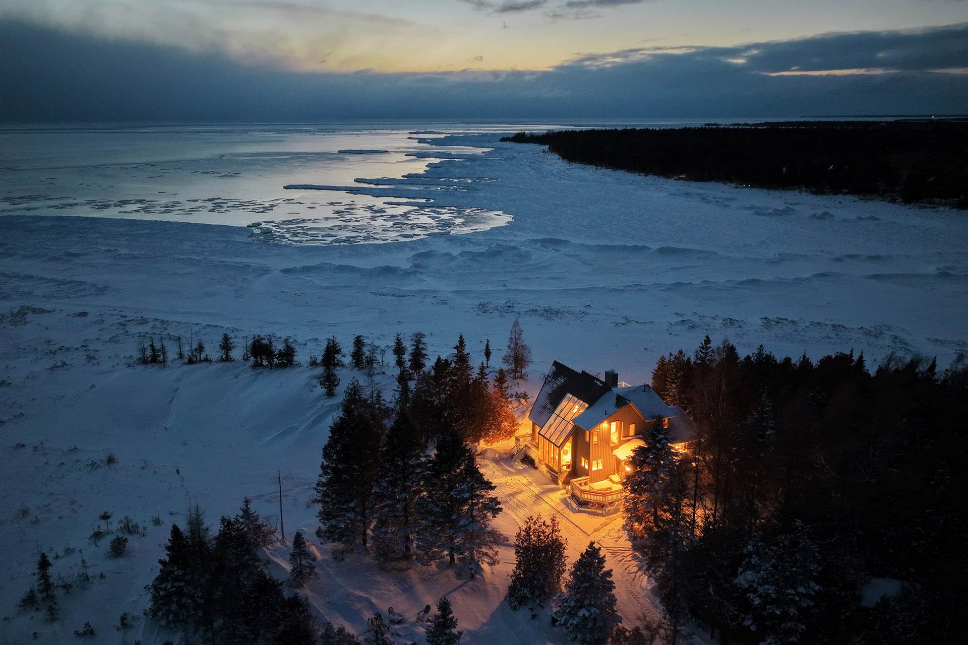 Winter dusk over the frozen bay, one warmly lit cottage among snowy trees
