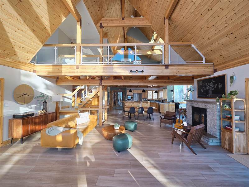 Open-concept great room with vaulted pine ceiling, loft above, leather sofa, brick fireplace and kitchen beyond