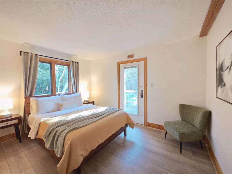 Bedroom with wood headboard, tan and sage bedding, green accent chair and glass door to the deck