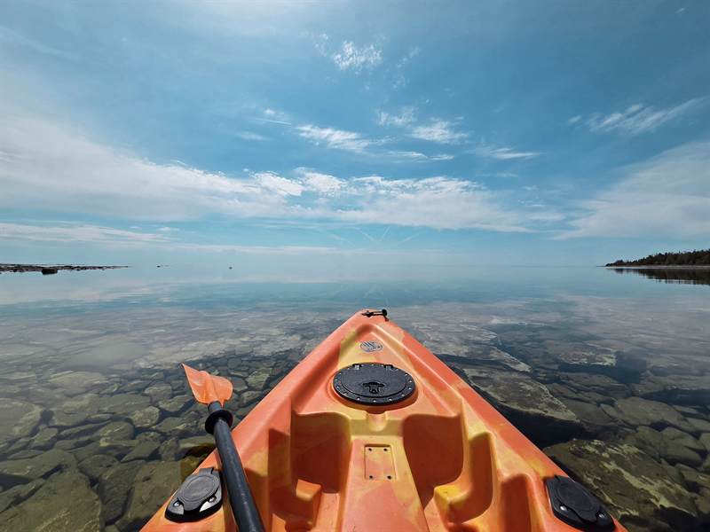 Bow of an orange kayak on still, clear water over cobblestones with sky reflected at the horizon