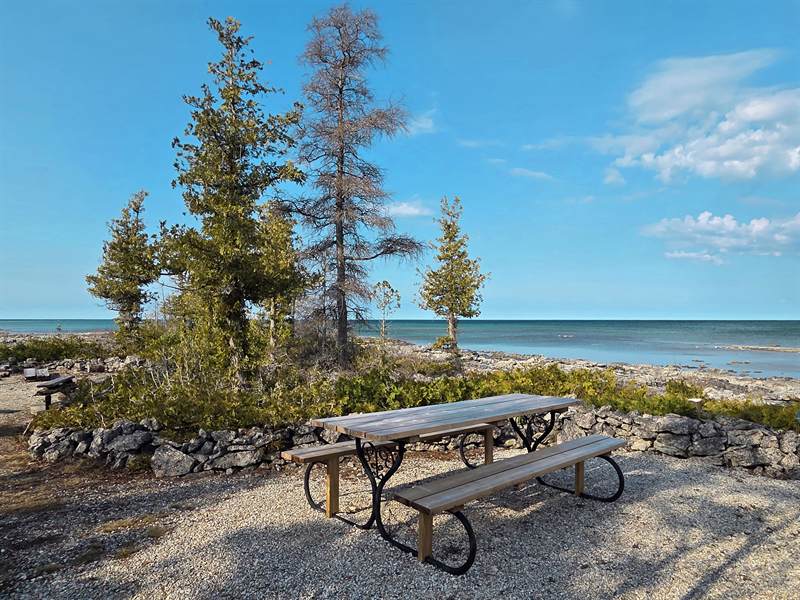 Wooden picnic table with iron legs on gravel beside a low stone wall, cedars and turquoise lake behind