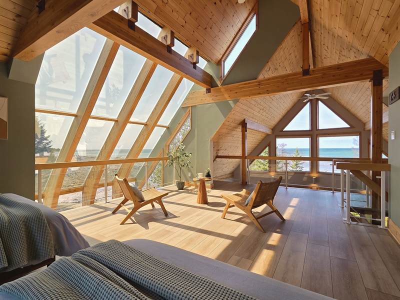 View from a bed across the loft with rattan chairs, angled glass wall and lake-view gable windows