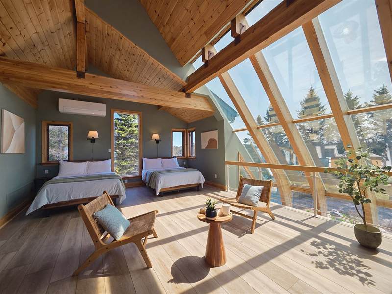 Loft bedroom with two white-dressed beds, wall sconces, rattan chairs and sloping glass wall