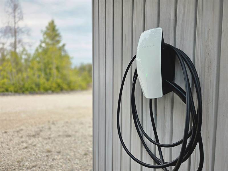 Tesla wall charger with coiled cable mounted on grey siding beside the gravel driveway