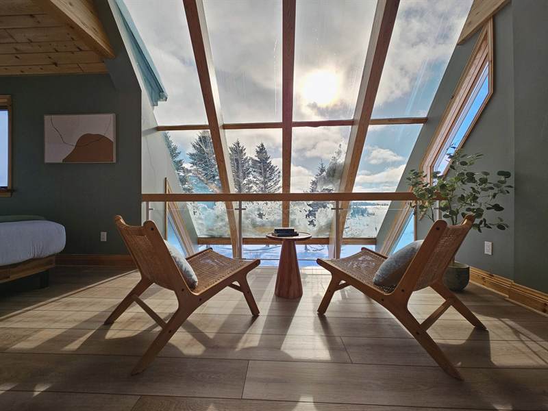 Two rattan lounge chairs and a side table facing a sloped glass wall with sun over snowy trees