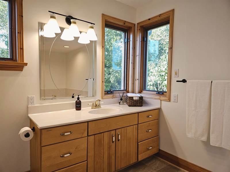 Bathroom vanity with wood cabinets, large mirror, sconce lights and corner windows onto greenery