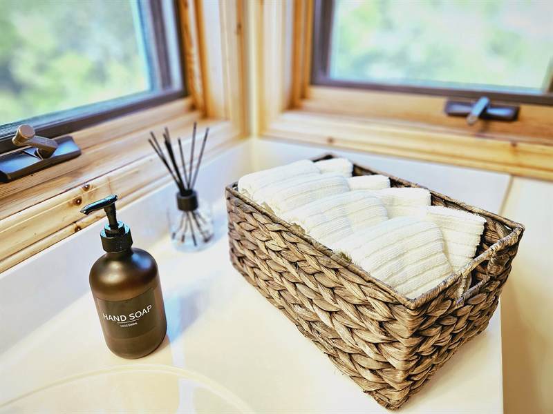 Wicker basket of rolled washcloths, hand soap and reed diffuser on a bathroom counter by corner windows